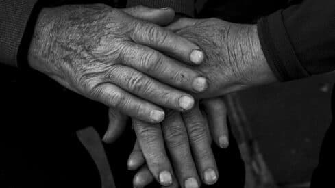 Two elderly pairs of hands in comforting gesture, black and white, showing wrinkles and texture.