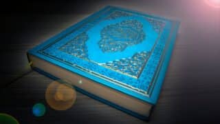 holy Quran with ornate blue cover and gold patterns on dark surface with light flares.