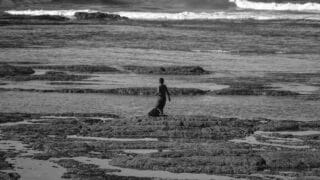 Person on rocky shore, facing ocean waves, black and white scene.