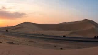 Desert scene with sand dunes, twilight sky, road, vehicles, and sparse vegetation.