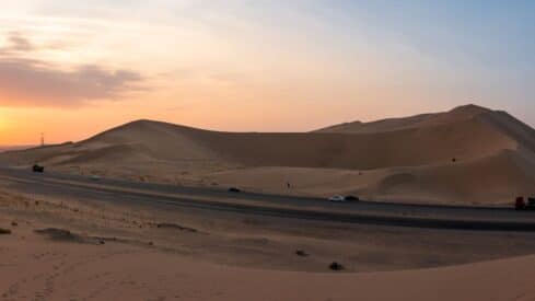 Desert scene with sand dunes, twilight sky, road, vehicles, and sparse vegetation.