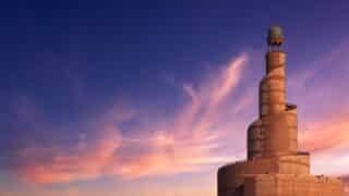 Spiral mosque minaret with geometric patterns at sunset, blue and pink sky, green dome on top.