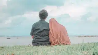 Two people sit on grass facing water under cloudy sky, one in a dark shirt, the other in brown garment.