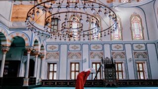 Interior of a mosque with ornate arches, stained glass, chandelier, person bowing in prayer.