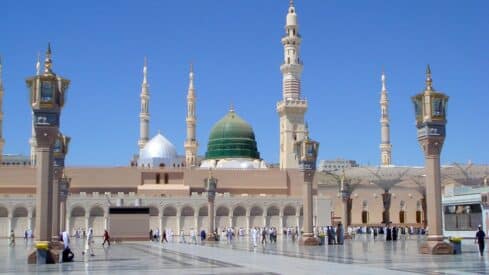 The Prophet's Mosque in Medina is shown with its green dome and minarets in the middle of a blue sky and people in the square.