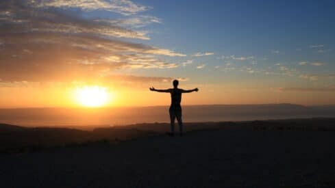 Person with outstretched arms on hilltop, silhouetted by sunrise or sunset, cloudy sky above.