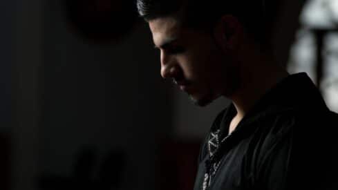 Young man in profile, dimly lit, wearing black shirt, contemplative expression with shadows.