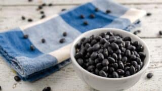 White bowl of black beans on wood, with blue checkered cloth and scattered beans nearby.