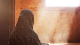 Person seated indoors with head covered by fabric, near sunlit window in dusty room.