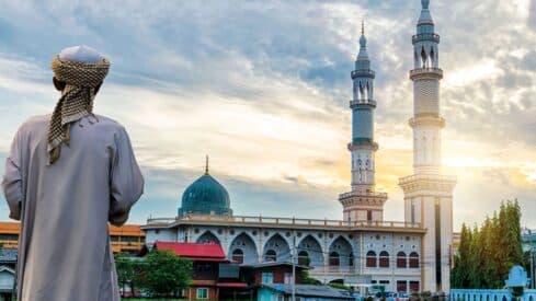 Person in traditional attire facing mosque with dome, two minarets, and an overcast sky.