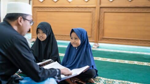 Man in white hat teaches two girls in abayas on a patterned carpet with a wooden backdrop. learning Quran