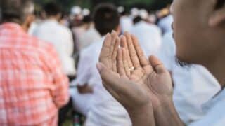 Person praying outdoors, hands together, with a crowd in white clothing visible in the background.