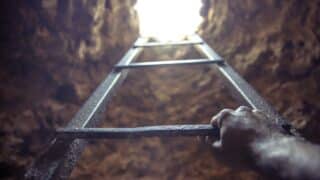 Person climbing metal ladder from rocky underground, light visible above.