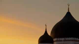 Two mosque domes with crescent moons silhouetted against a sunset-colored sky.