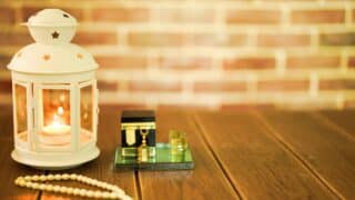 White lantern with candle, black cube box, prayer beads on wood table, brick wall backdrop.