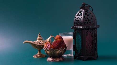 Decorative setup with a brass oil lamp, dates, milk, and a lantern against a teal background.