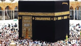 Kaaba draped in black cloth with golden calligraphy, surrounded by a crowd in a mosque.