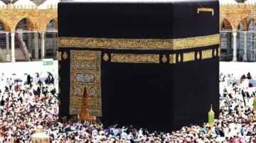 Kaaba draped in black cloth with golden calligraphy, surrounded by a crowd in a mosque.