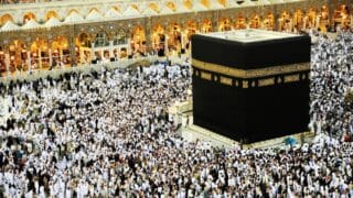 Crowd gathers around the Kaaba in Masjid al-Haram, under bright lights, during pilgrimage.