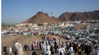 A crowd in traditional white attire with colorful umbrellas in hajj, mountains, buildings, and vehicles.