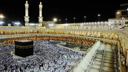 Night view of Kaaba in Masjid al-Haram, Mecca, surrounded by a large crowd and lit minarets.