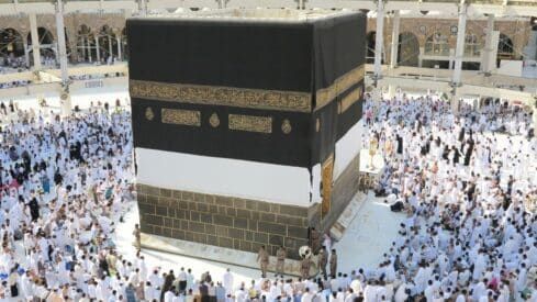 Crowd gathered around the Kaaba in Mecca, Saudi Arabia, with surrounding architecture visible.