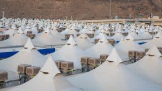 Large white tents with pointed tops and AC units, rocky hillside background.