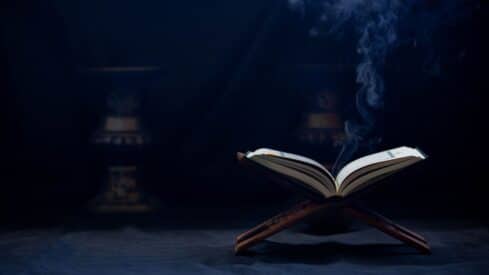 Open Quran on a wooden stand with rising smoke against a dark background.