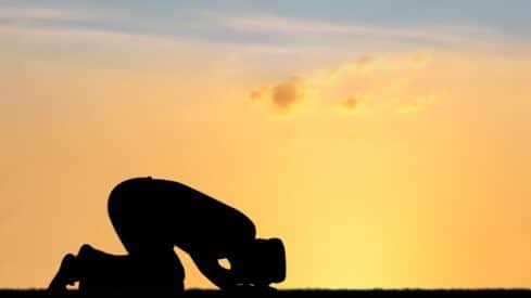 Silhouette of a kneeling person praying on grass against an orange sunset sky.