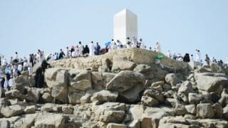 People gathered on a rocky hill around a tall monument Arafat , with some using umbrellas for shade.