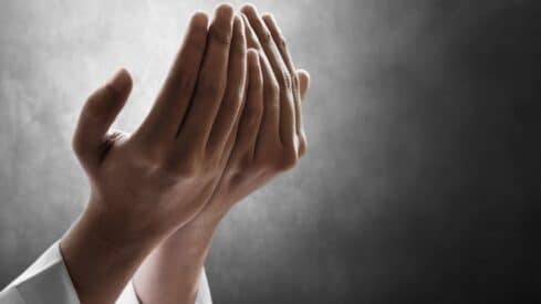 Hands raised, palms open in prayer or supplication, against a dark gray background.