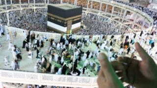 Large crowd gathered around the Kaaba in Mecca, with pilgrims in traditional attire.