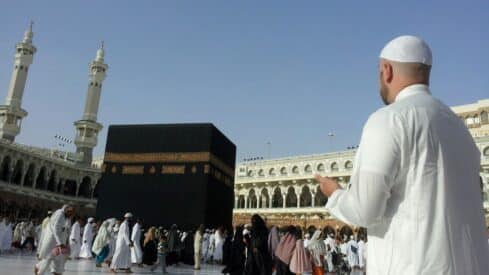 Man in reverence near Kaaba, surrounded by people, with two minarets at Masjid al-Haram.