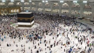 Large crowd surrounds Kaaba inside Grand Mosque, Mecca, with arches and multi-levels visible.