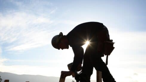 Silhouette of a construction worker in hard hat bending, sun shining behind, against bright sky.