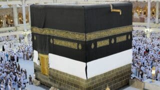 Kaaba draped in black with gold text, surrounded by people in white in a mosque.