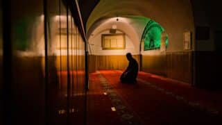 Person kneels in prayer on carpet in dimly lit room with arched ceiling and wood-paneled walls.