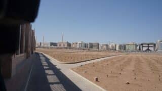 Dirt field with markers, bordered by walkways; modern buildings and minarets in the background.