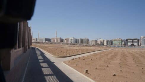 Dirt field with markers, bordered by walkways; modern buildings and minarets in the background.