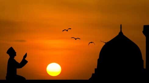 Silhouette of a praying person at sunset, birds flying, dome structure on the right.