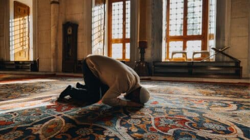 Person kneels in prayer on a colorful carpet in a sunlit room with large windows and columns.