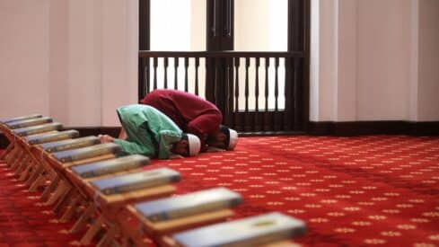 Two people pray on a red carpet near book stands in a room with a large window.