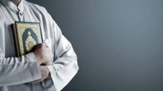 Person in robe holding holy Quran against plain background.