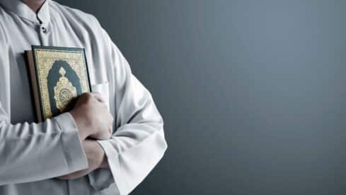 Person in robe holding holy Quran against plain background.