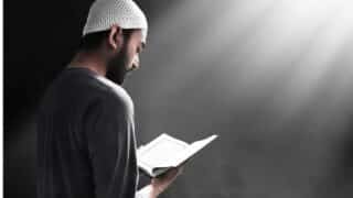 Man in white cap reading Quran under overhead light in dim setting.