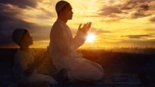 Man and child in traditional attire kneel and pray at sunset near cityscape.