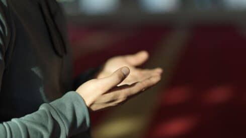 Person in dark garment with hands open in prayer gesture; warm, blurred background.