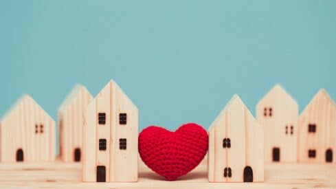 Wooden house models in a row with red knitted heart between two, on blue background.