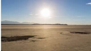 Vast desert landscape under a clear blue sky with sun and distant mountains on the horizon.