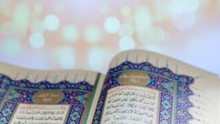 Open Quran with Arabic text, ornate blue and red borders, blurred light background.